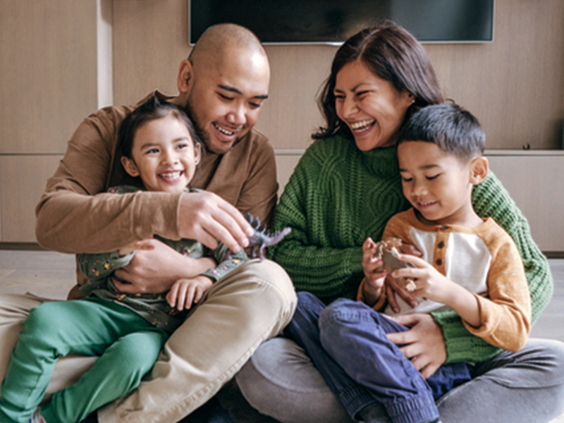 Mom and dad with their two young children in their living room, playing with plastic dinosaurs.