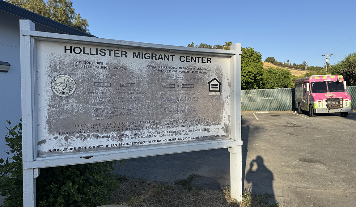 Sign reading "Hollister Migrant Center" in front of a building and parking lot, where a colorful van is parked.