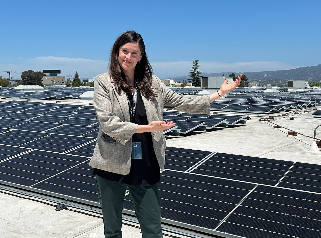Director Kerry Fleisher stands in front of a rooftop solar array, gesturing toward the panels.