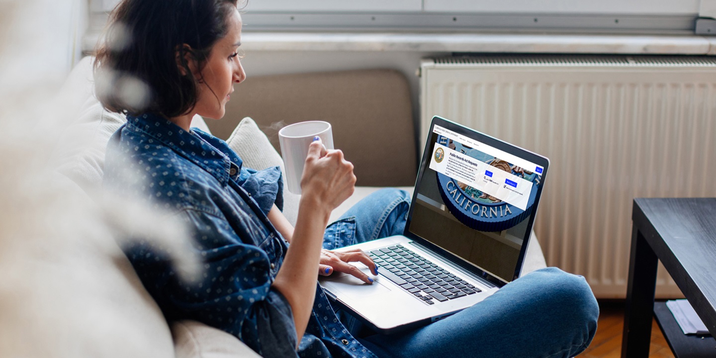 A woman sits cross-legged on her couch, holding a cup of coffee and looking at the PRA webpage on the laptop in her lap.